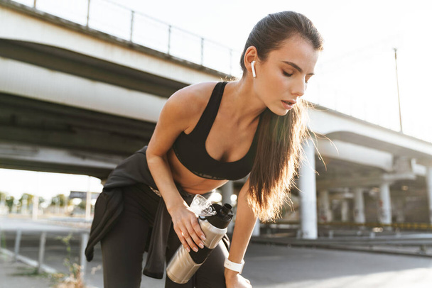Image of a serious sports fitness woman outdoors listening music with earphones drinking water. - Foto, Imagen