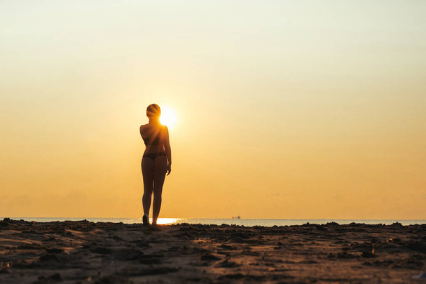 Silhouette of woman in a swimsuit standing on the beach opposite sea and rising sun. Summer and freedom concept. Sun rays (beam) shining through female legs. - Foto, Imagen