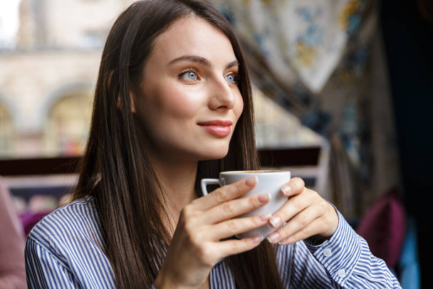 Close up of a beautiful young brunette woman drinking coffee while sitting in the cafe indoors - Foto, afbeelding