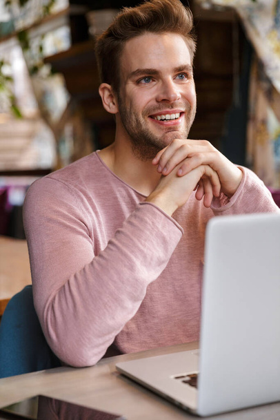 Bonito sorridente jovem trabalhando no computador portátil enquanto sentado no café
 - Foto, Imagem