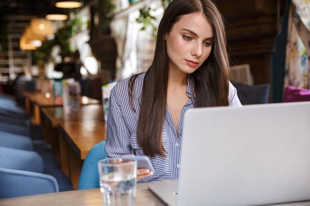 Ελκυστική σκεπτική νεαρή μελαχρινή γυναίκα που εργάζεται σε φορητό υπολογιστή, ενώ κάθεται στο καφέ σε εσωτερικούς χώρους - Φωτογραφία, εικόνα