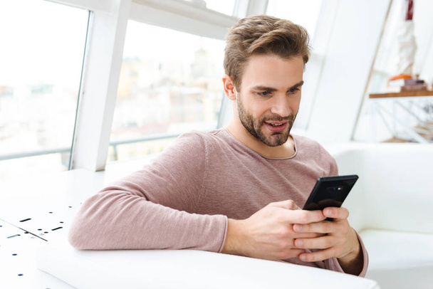 Image of young bearded man using smartphone while sitting on sofa by window in cafe indoors - 写真・画像