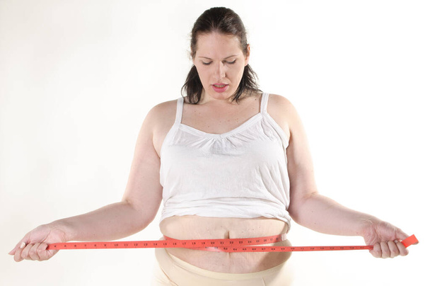 Adult overweight woman with black hair wears white underwear and measures her abdominal circumference with a tape measure. She is frustrated and sad,against a white background. - Photo, image