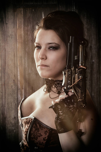 a woman in steam punk outfit holding a gun and looking into the camera in front of a wooden background - Photo, image