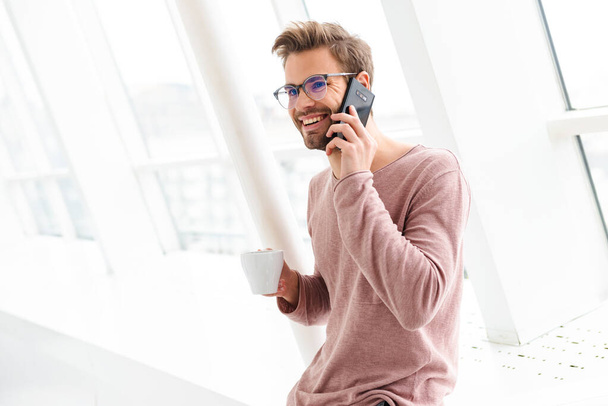 Image of young bearded man wearing eyeglasses talking on smartphone while standing over bright window indoors - Foto, Imagem