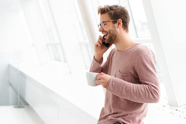 Image of young bearded man wearing eyeglasses talking on smartphone while standing over bright window indoors - Foto, Bild