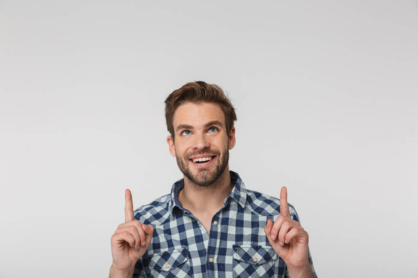 Portrait of joyful young man wearing plaid shirt smiling and pointing fingers upward isolated over white background - 写真・画像
