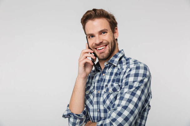 Portrait of joyful young man wearing plaid shirt smiling and talking on cellphone isolated over white background - Fotoğraf, Görsel