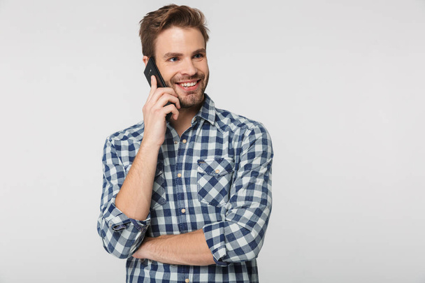 Portrait of joyful young man wearing plaid shirt smiling and talking on cellphone isolated over white background - Photo, image