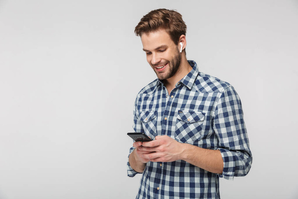 Portrait of happy young man wearing plaid shirt using cellphone and earphones isolated over white background - Foto, imagen