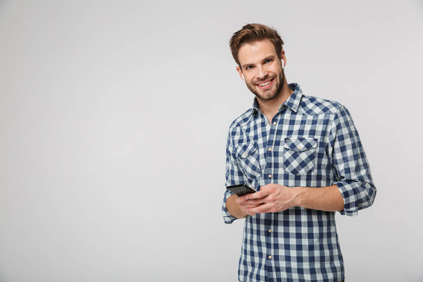 Portrait of happy young man wearing plaid shirt using cellphone and earphones isolated over white background - Foto, Bild