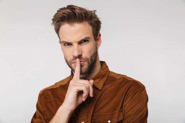 Portrait of displeased young man wearing brown shirt gesturing silence sign isolated over white background - 写真・画像