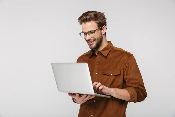 Portrait of cheerful young man wearing eyeglasses using laptop and smiling isolated over white background - Φωτογραφία, εικόνα