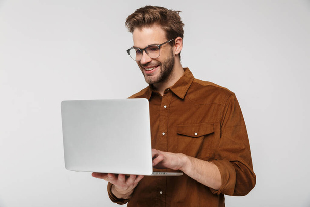 Portrait of cheerful young man wearing eyeglasses using laptop and smiling isolated over white background - Fotografie, Obrázek