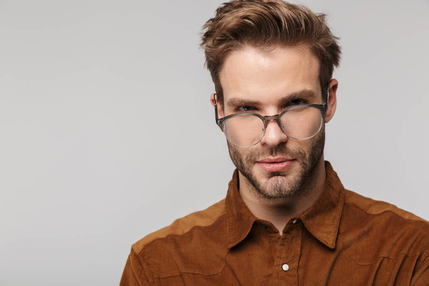 Portrait of unshaven young man wearing eyeglasses posing and looking at camera isolated over white background - Foto, afbeelding
