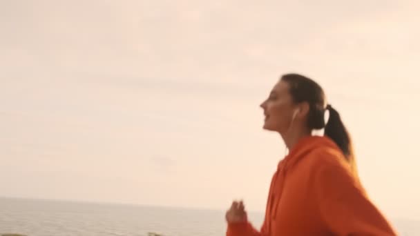 Πλευρική άποψη του Happy όμορφη αθλητική γυναίκα σε ακουστικά τρέχει και κοιτάζοντας μακριά στην παραλία - Πλάνα, βίντεο