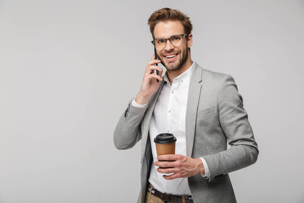 Portrait of smiling handsome man in eyeglasses talking on cellphone and holding paper cup isolated over white background - Valokuva, kuva
