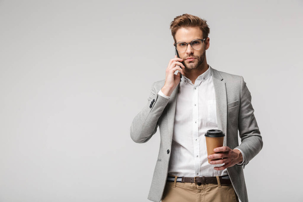 Portrait of serious handsome man in eyeglasses talking on cellphone and holding paper cup isolated over white background - Foto, Bild