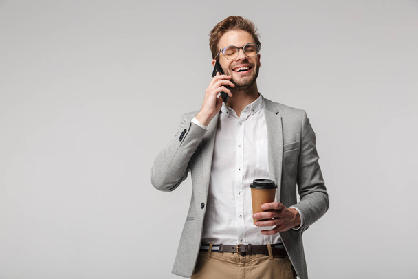Portrait of smiling handsome man in eyeglasses talking on cellphone and holding paper cup isolated over white background - Foto, afbeelding