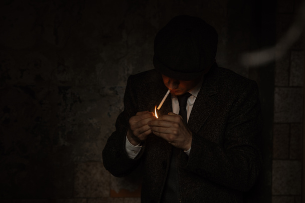 A man smokes a cigarette in the image of an English retro gangster of the 1920s dressed in a coat, suit and flat cap in Peaky blinders style. - Фото, изображение