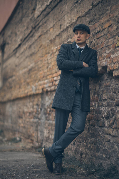 A man posing in the image of an English retro gangster of the 1920s dressed in Peaky blinders style near old brick wall. - Foto, afbeelding