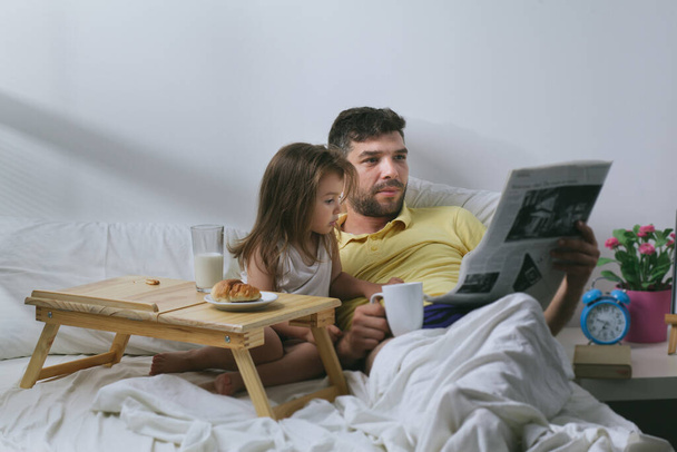 Father and daughter having breakfast in bed in the morning. Father reading newspaper - Fotografie, Obrázek