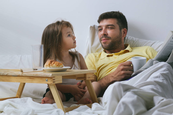 Father and daughter having breakfast in bed in the morning. Father reading newspaper - Foto, immagini