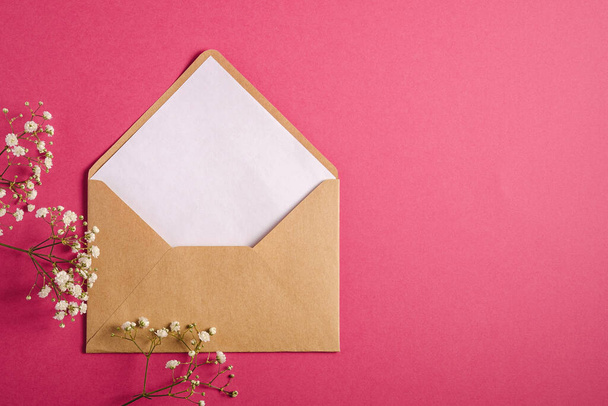 Sobre de papel marrón Kraft con tarjeta vacía blanca, flores de gypsophila, fondo rosa rojo, plantilla de maqueta
 - Foto, Imagen