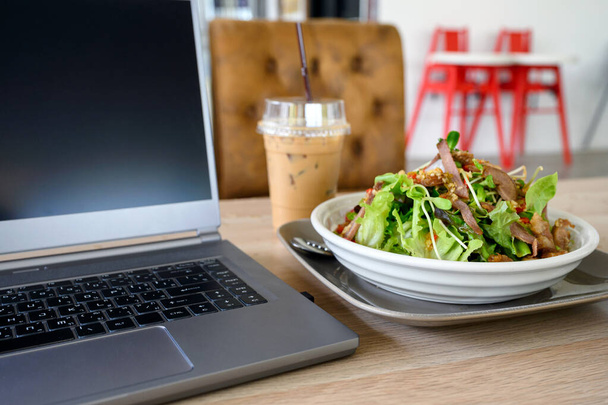 Moderní laptop s pikantním vepřovým salátem a ledovou kávou na dřevěném stole - Fotografie, Obrázek