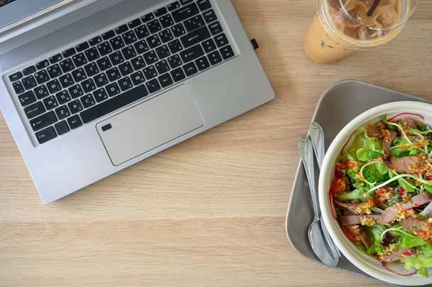 Pose plate, ordinateur portable moderne avec salade de porc épicée et café glacé sur table en bois
 - Photo, image