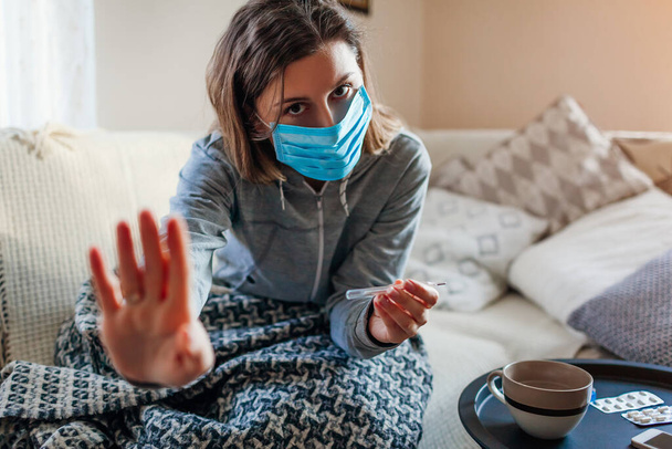 Ill woman checks thermometer having fever. Girl wears protective mask having cold flu takes pills at home shows stop sign warning about disease. Healthcare - Photo, Image