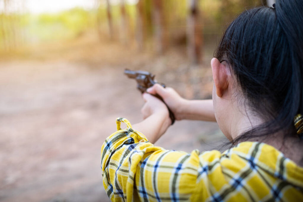 Portrait the farmer asea woman wearing a yellow Shirt at the shooting shot from old revolver gun in the farm, Young girl sitting in the attitude of aiming and looking through the sight handgun - 写真・画像