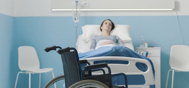 Paciente hospitalizado deitado na cama e cadeira de rodas em primeiro plano, tratamento médico e conceito de lesão
 - Foto, Imagem