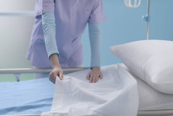 Η έμπειρη νεαρή νοσοκόμα που εργάζεται στο νοσοκομείο, στρώνει το κρεβάτι και αλλάζει σεντόνια, υπηρεσίες υγείας και βοήθεια έννοια - Φωτογραφία, εικόνα