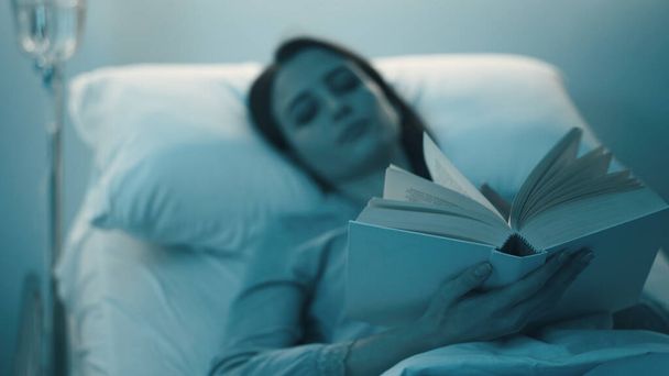 Young sleepless woman lying in the hospital bed at night and reading a book - Фото, изображение