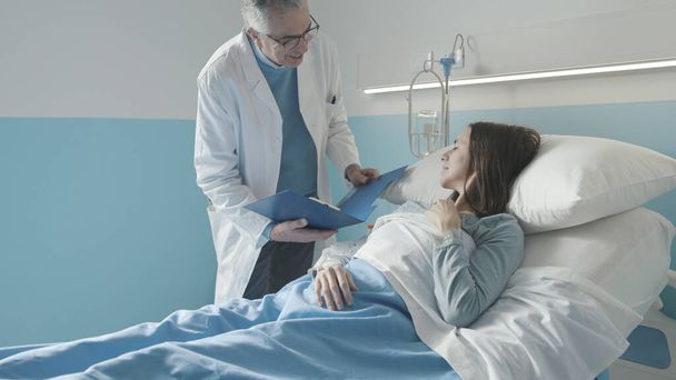 Professional doctor visiting a female patient lying in bed at the hospital, he is checking her medical records - Foto, Imagem