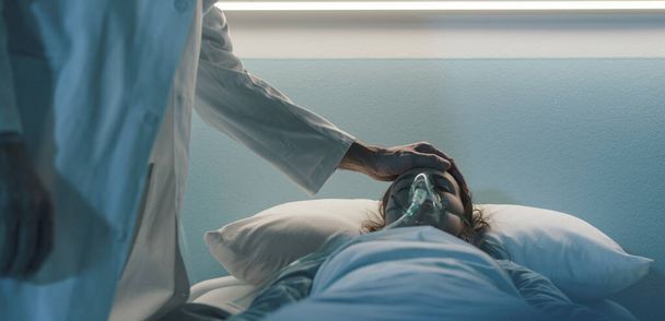 Doctor assisting a hospitalized patient at night and touching her forehead: she is lying in bed with an oxygen mask - Φωτογραφία, εικόνα