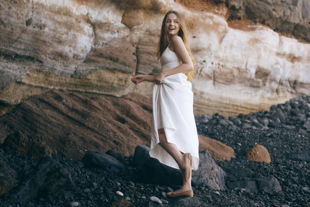Gorgeous girl in a long white dress on the ocean at the beach. The long-haired girl is inherited by nature, mountains, stones, the beach. Girl on vacation, light dress, summer, date. Bride - Foto, Imagem