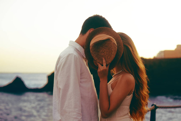 夫婦は海と山の背景にキスをします。ロマンチックな写真、女の子は帽子、キスで覆われています。トーンのスタイリッシュな写真。美しい藁帽子。旅行はロマンチックです. - 写真・画像