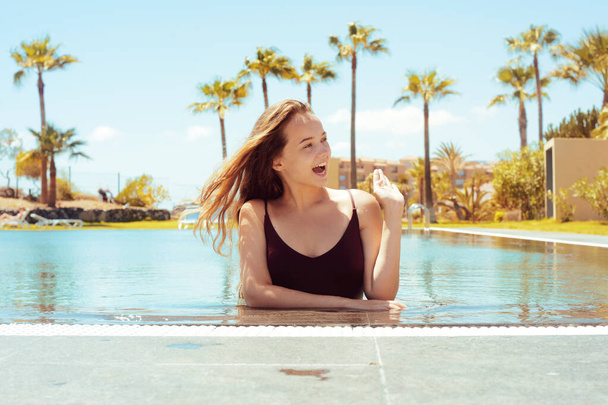 Happy girl in the pool, blonde with long hair in a red bathing suit. The girl rejoices in traveling, flirts, smiles, rejoices. Traveling. Girl on a background of palm trees. - Foto, imagen