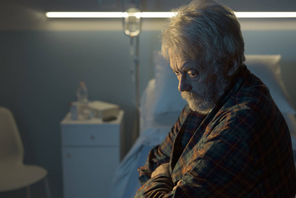 Depressed senior man sitting on the hospital bed alone at night, he feels lonely and abandoned - Фото, зображення