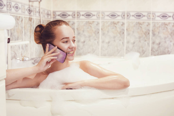 The girl is lying in the whirlpool with foam and talking on a waterproof phone. The girl relaxes and gossips. Poetz girl in the foam. Conversation with a girlfriend. - Photo, Image