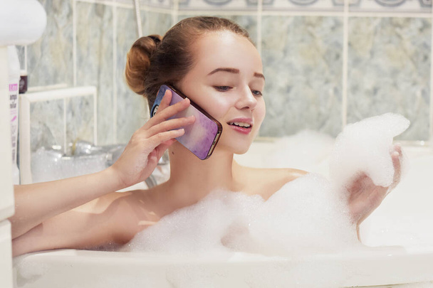 The girl is lying in the whirlpool with foam and talking on a waterproof phone. The girl relaxes and gossips. Poetz girl in the foam. Conversation with a girlfriend. - Photo, Image