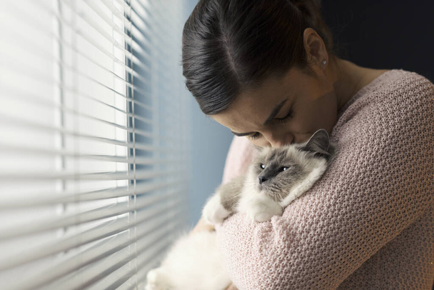 Relaxed woman standing next to a window, she is holding and kissing her beautiful cat - Foto, afbeelding