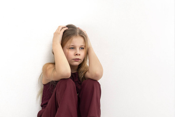 Shot of a young girl wearing a burgundy jumpsuit siting on the floor by the wall and thinks or sad about something - Foto, afbeelding