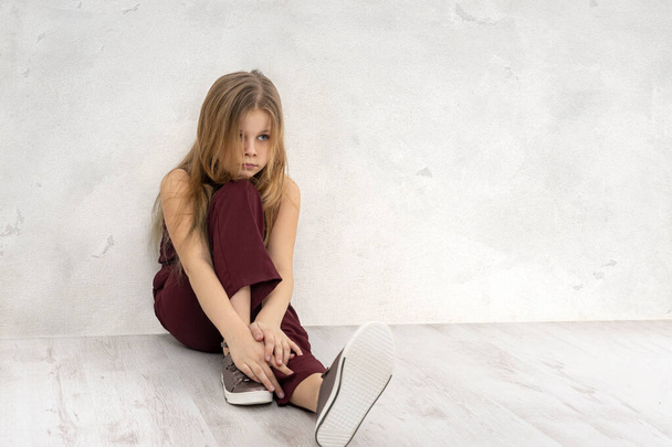 Shot of a young girl wearing a burgundy jumpsuit siting on the floor by the texture wall and thinks or sad about something - Foto, Imagen
