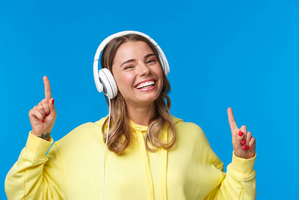 Music, lifestyle and youth concept. Close-up portrait of joyful smiling blond girl listening songs in headphones, dancing and move fingers rhythm, standing happy over blue background - Valokuva, kuva