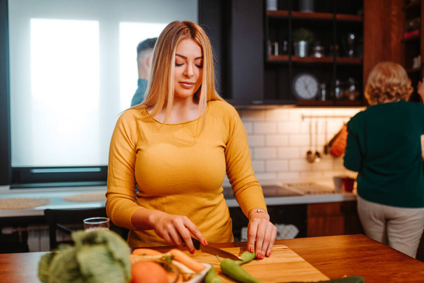 Young blonde woman making salad in kitchen. She is chopping vegetables with a kitchen knife. - Foto, Bild