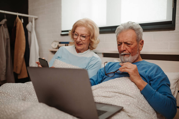 Eine ältere blonde Frau mit Brille zeigt ihrem Mann, der neben ihr im Bett liegt und den Laptop auf seinem Schoß hält, etwas auf ihrem Smartphone. Älteres Ehepaar fotografiert. - Foto, Bild
