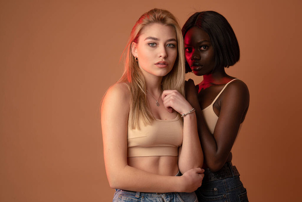 Dos chicas internacionales en sujetador beige, chicas africanas y europeas mirando a la cámara aisladas sobre fondo naranja oscuro
 - Foto, imagen